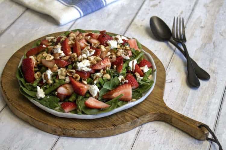 A round rustic white serving platter with strawberry asparagus salad with ginger balsamic dressing is displayed in the center of the picture; the plate is sitting on a round wooden paddle, a black spoon and fork are to the right of it and a white and blue napkin is in the back - all items are displayed on a white wooden surface