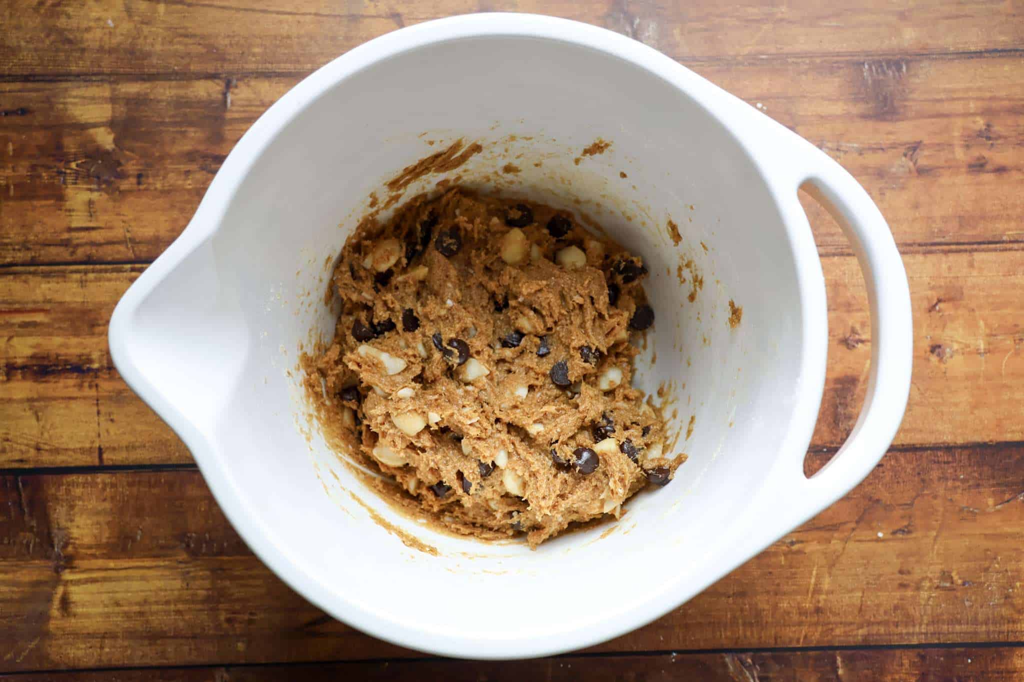 the dough, including chocolate chips and chopped macadamia nuts, for delicious gluten-free chocolate chip macadamia cookies is displayed in a white bowl, which is placed on a wooden surface