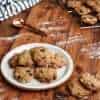 A plate of delicious gluten free chocolate chip macadamia cookies is in the center of the picture on a wooden surface with two additional cookies next to the plate; a cooling rack with additional cookies is in the background, along with a white and grey striped napkin and a copper colored measuring spoon and a little bit of flour is scattered across the wooden surface