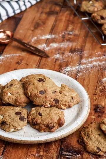 A plate of delicious gluten free chocolate chip macadamia cookies is in the center of the picture on a wooden surface with two additional cookies next to the plate; a cooling rack with additional cookies is in the background, along with a white and grey striped napkin and a copper colored measuring spoon and a little bit of flour is scattered across the wooden surface