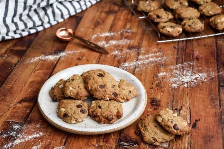 A plate of delicious gluten free chocolate chip macadamia cookies is in the center of the picture on a wooden surface with two additional cookies next to the plate; a cooling rack with additional cookies is in the background, along with a white and grey striped napkin and a copper colored measuring spoon and a little bit of flour is scattered across the wooden surface