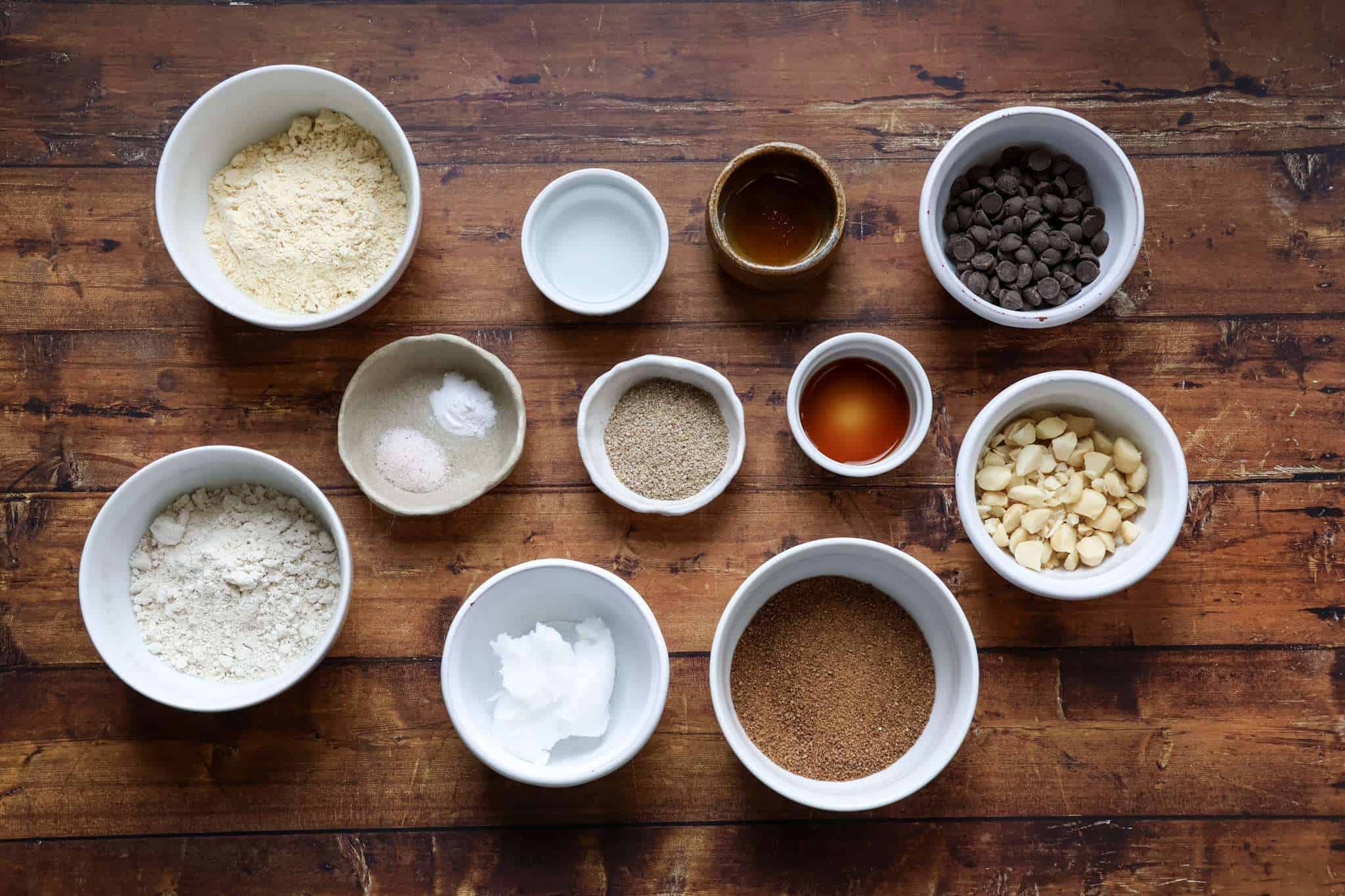 The ingredients for delicious gluten free chocolate chip macadamia cookies are displayed in bowls on a wooden surface, including chickpea flour, oat flour, coconut sugar, sea salt, baking soda, water, ground chia, vanilla extract, maple syrup, coconut oil, chocolate chips, and macadamia nuts