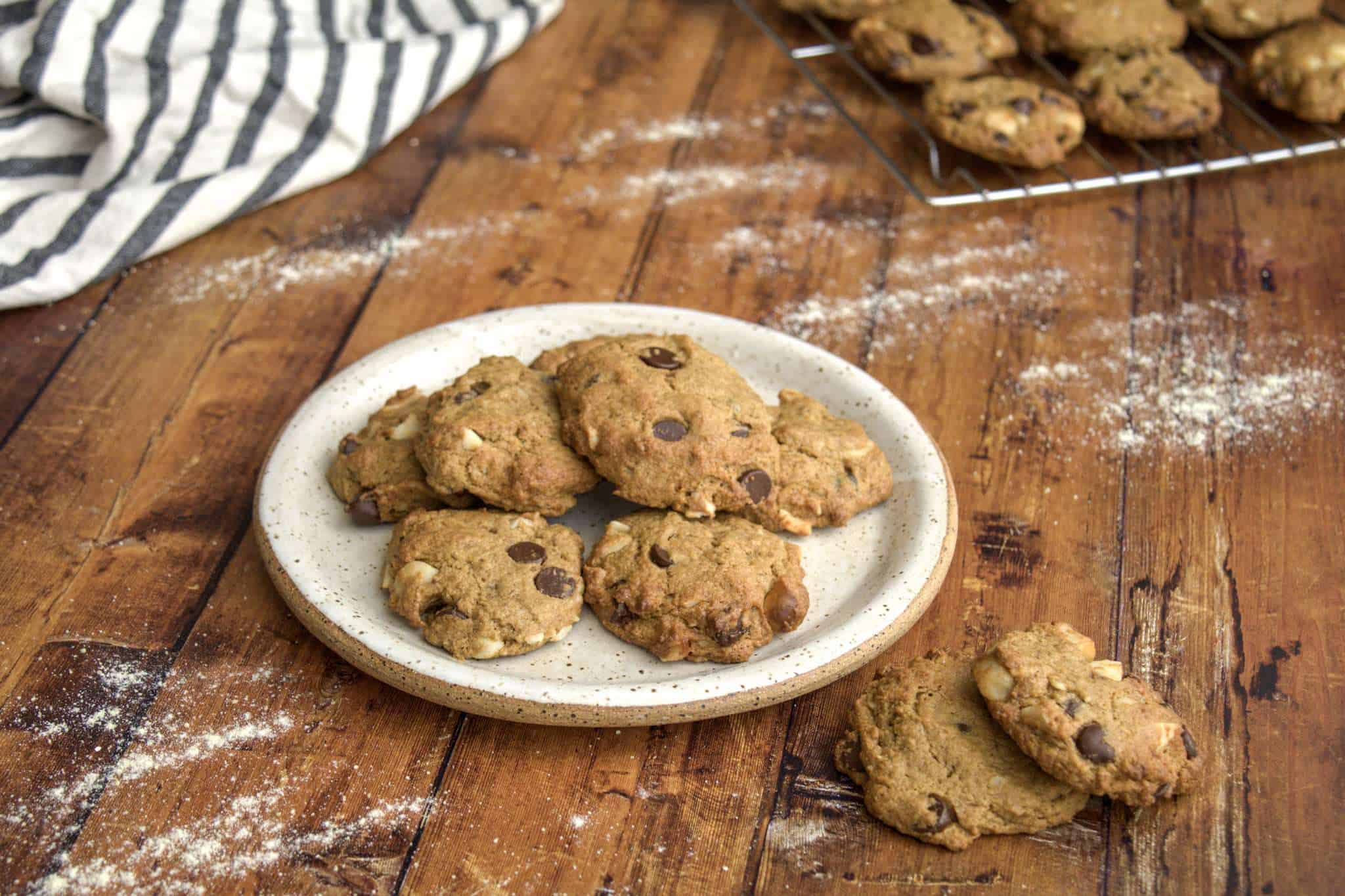 A plate of delicious gluten free chocolate chip macadamia cookies is in the center of the picture on a wooden surface with two additional cookies next to the plate; a cooling rack with additional cookies is in the background, along with a white and grey striped napkin and a little bit of flour is scattered across the wooden surface