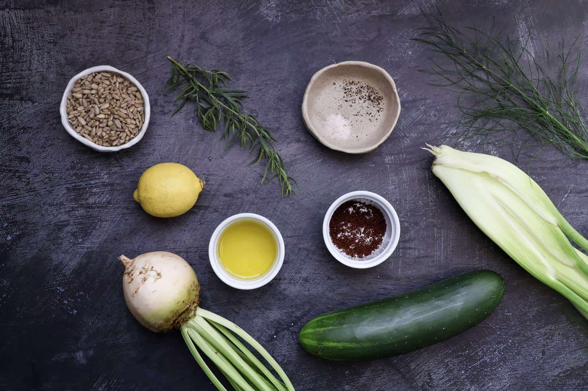 All ingredients for cucumber bites with sunflower beet and fennel puree are displayed on a dark grey concrete style surface- the ingredients include cucumber, a white beet, fennel bulb, sunflower seeds, tarragon sprig, fennel fronds, a lemon, extra virgin olive oil, sumac, fine sea salt, and pepper
