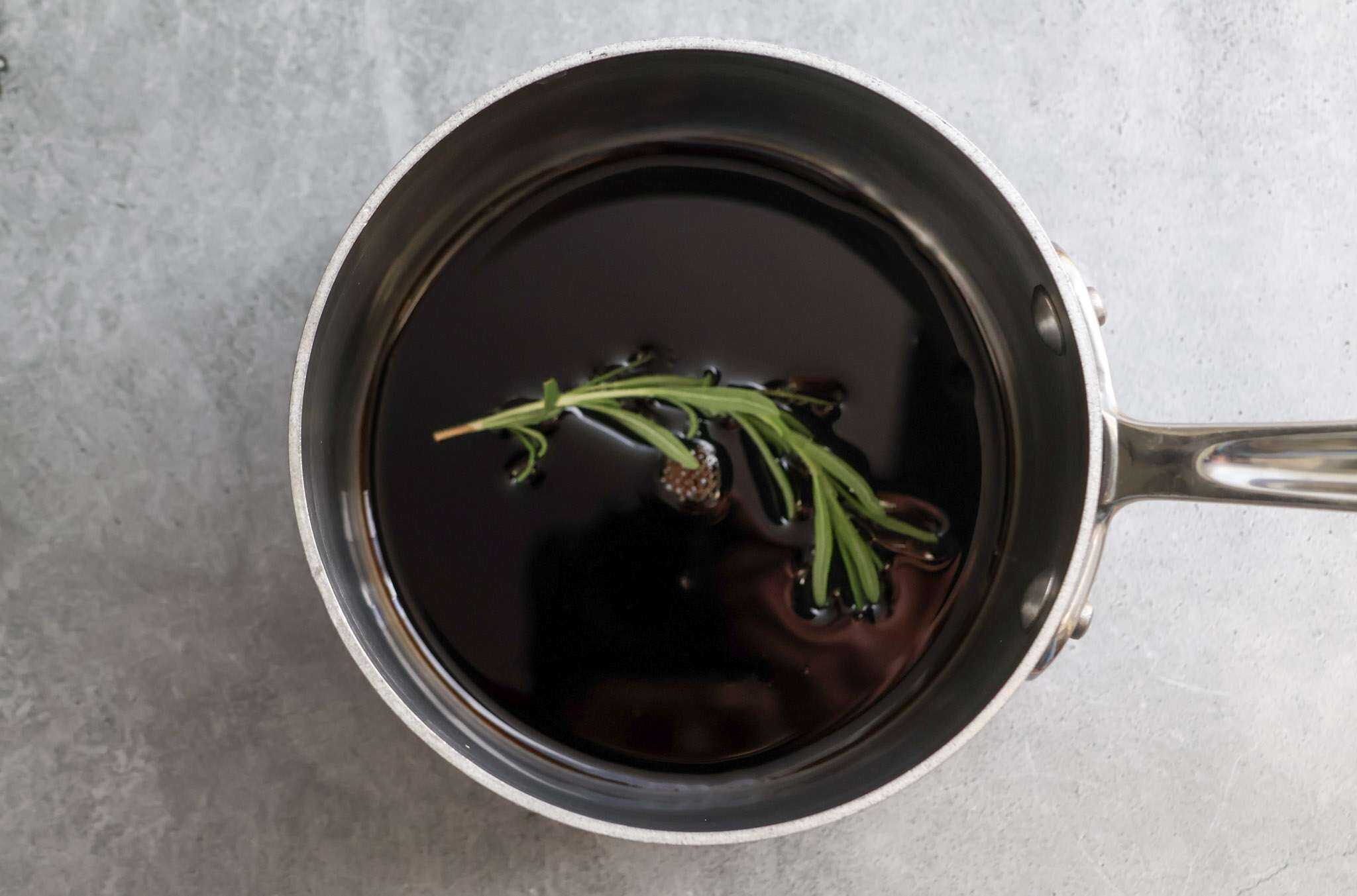 a small saucepan is displayed on a grey concrete style background - in the bowl is balsamic vinegar with a rosemary sprig