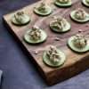 Cucumber bites with sunflower, beet, and fennel purée, garnished with sunflower seeds, fennel fronds, and sumac on a rustic wooden board with a striped napkin
