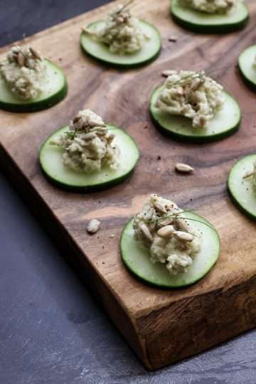Cucumber bites with sunflower, beet, and fennel purée, garnished with sunflower seeds, fennel fronds, and sumac on a rustic wooden board with a striped napkin