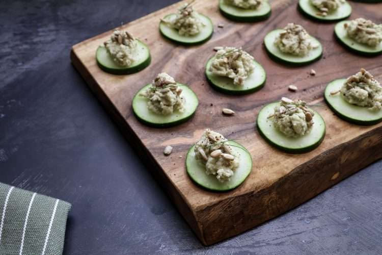 Cucumber bites with sunflower, beet, and fennel purée, garnished with sunflower seeds, fennel fronds, and sumac on a rustic wooden board with a striped napkin