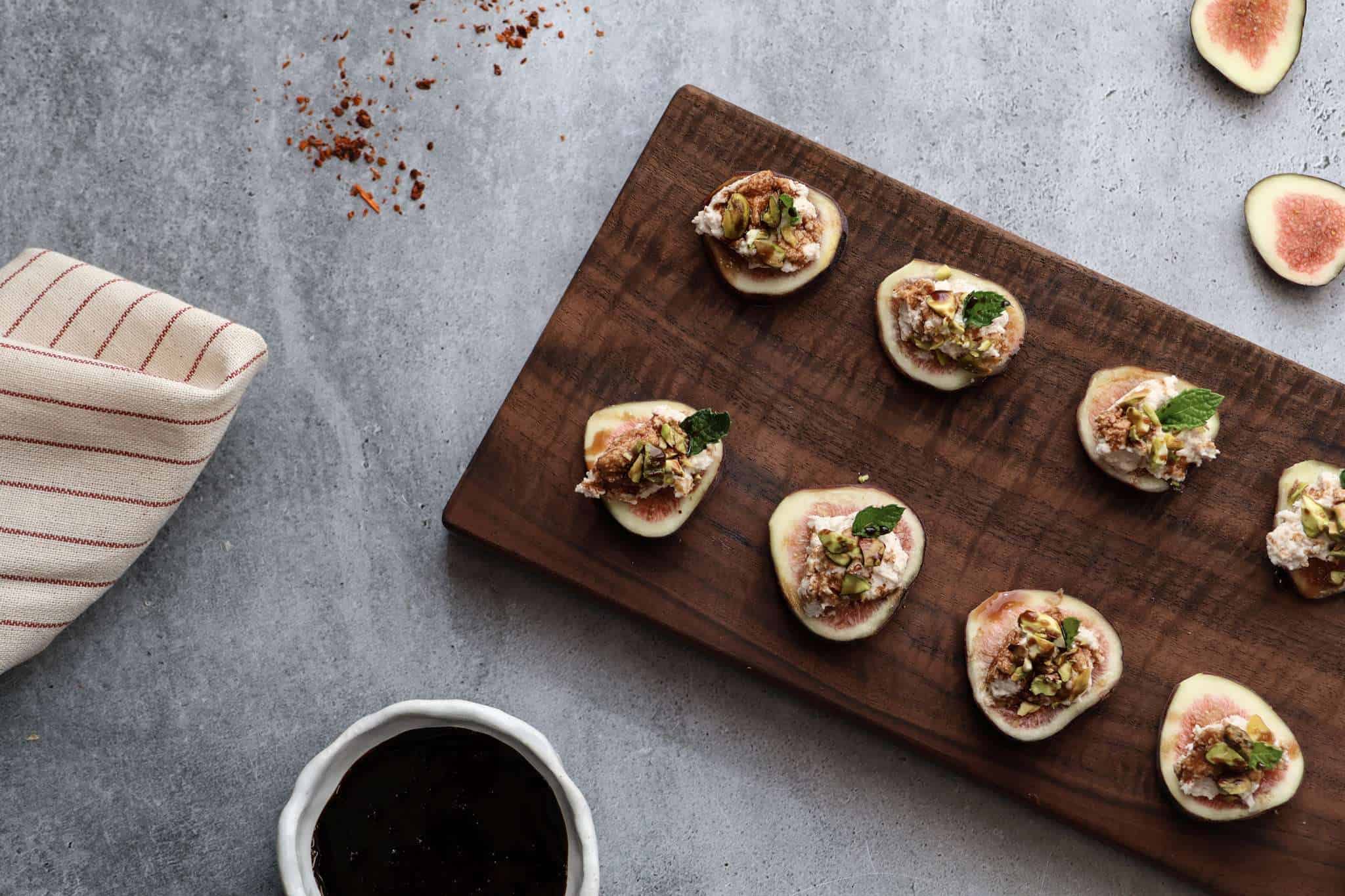A rectangular wooden board is displayed on a grey concrete style background-in the front of the picture is a small white bowl with balsamic reduction, to the left is a beige and red striped napkin, along the top is some scattered aleppo pepper and toward the top right are a couple little fig slices. On the board are slices of fig, topped with a spiced goat cheese mixture, topped with chopped pistachios, a small mint leaf and a drizzle of rosemary balsamic reduction