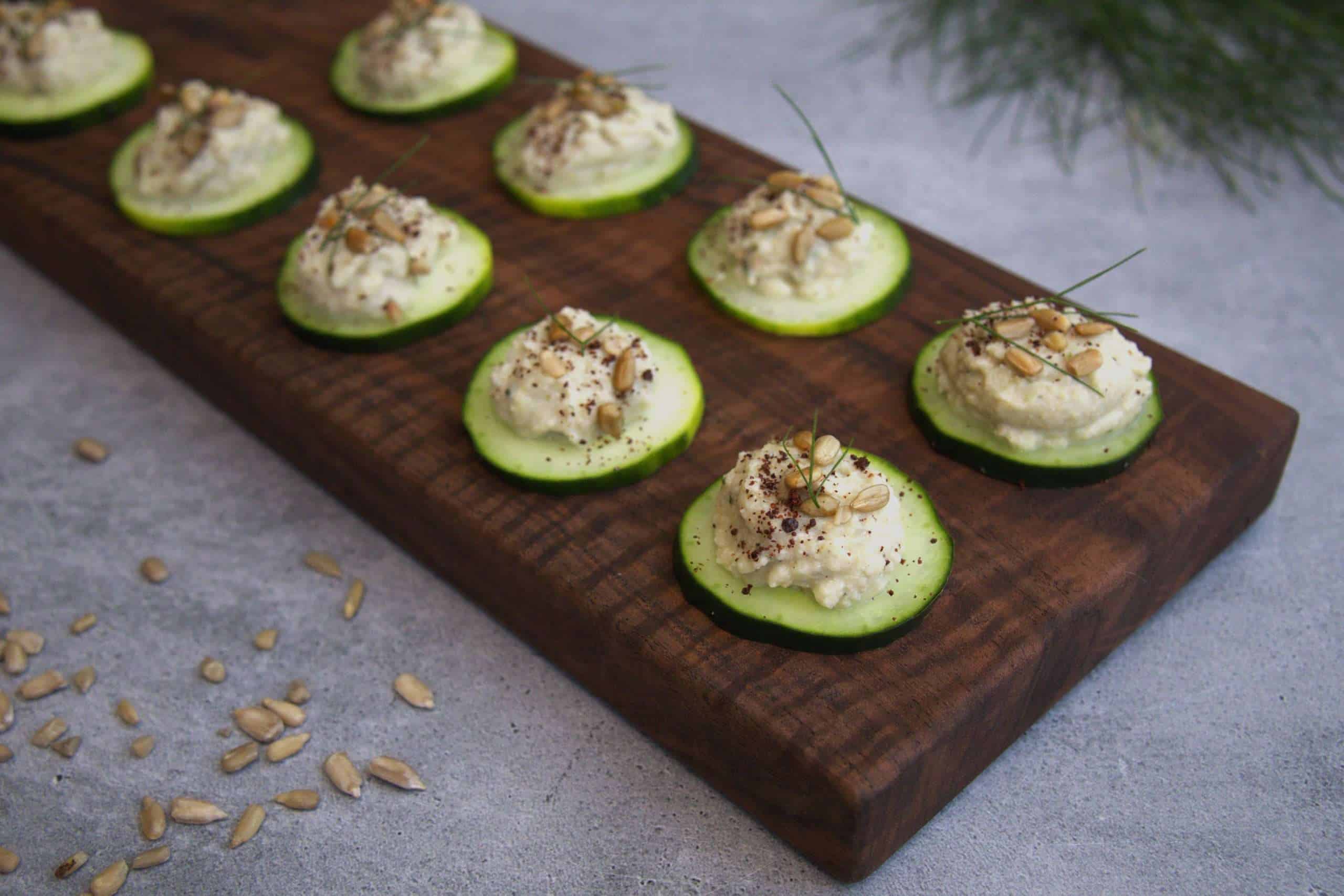 Cucumber bites with sunflower, beet, and fennel purée, garnished with sunflower seeds, fennel fronds, and sumac on a wooden board with sunflower seeds and fennel fronds scattered in the front and back of the picture