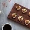 A rectangular wooden board is displayed on a grey concrete style background-in the front of the picture is a small white bowl with balsamic reduction, to the left is a beige and red striped napkin, along the top is some scattered aleppo pepper and toward the top right are a couple little fig slices. On the board are slices of fig, topped with a spiced goat cheese mixture, topped with chopped pistachios, a small mint leaf and a drizzle of rosemary balsamic reduction