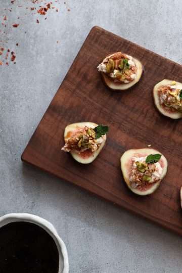A rectangular wooden board is displayed on a grey concrete style background-in the front of the picture is a small white bowl with balsamic reduction, to the left is a beige and red striped napkin, along the top is some scattered aleppo pepper and toward the top right are a couple little fig slices. On the board are slices of fig, topped with a spiced goat cheese mixture, topped with chopped pistachios, a small mint leaf and a drizzle of rosemary balsamic reduction