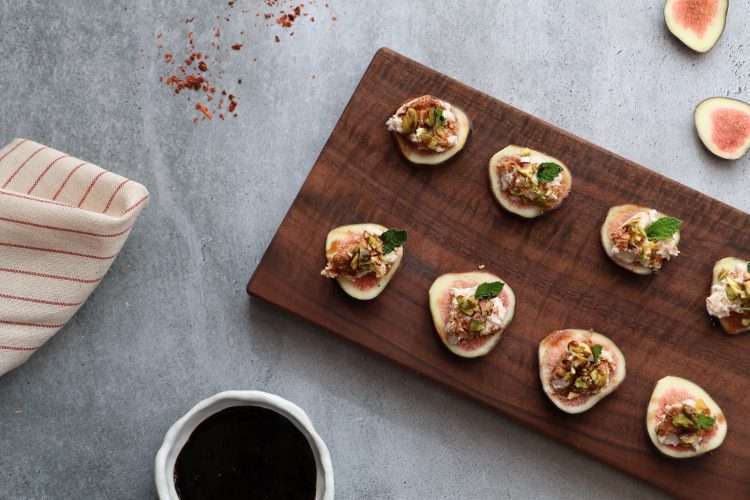 A rectangular wooden board is displayed on a grey concrete style background-in the front of the picture is a small white bowl with balsamic reduction, to the left is a beige and red striped napkin, along the top is some scattered aleppo pepper and toward the top right are a couple little fig slices. On the board are slices of fig, topped with a spiced goat cheese mixture, topped with chopped pistachios, a small mint leaf and a drizzle of rosemary balsamic reduction