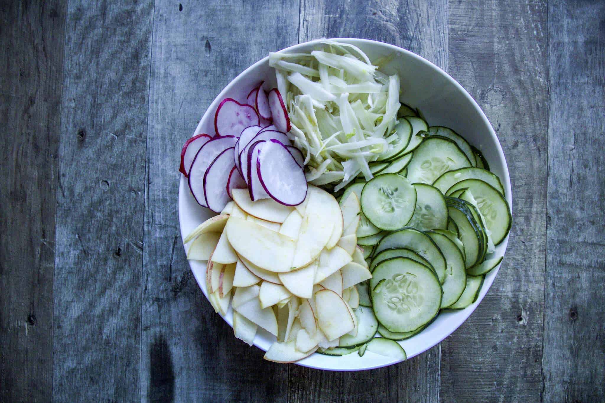 A white bowl containing thinly sliced, apple, cucumber, radish, and fennel