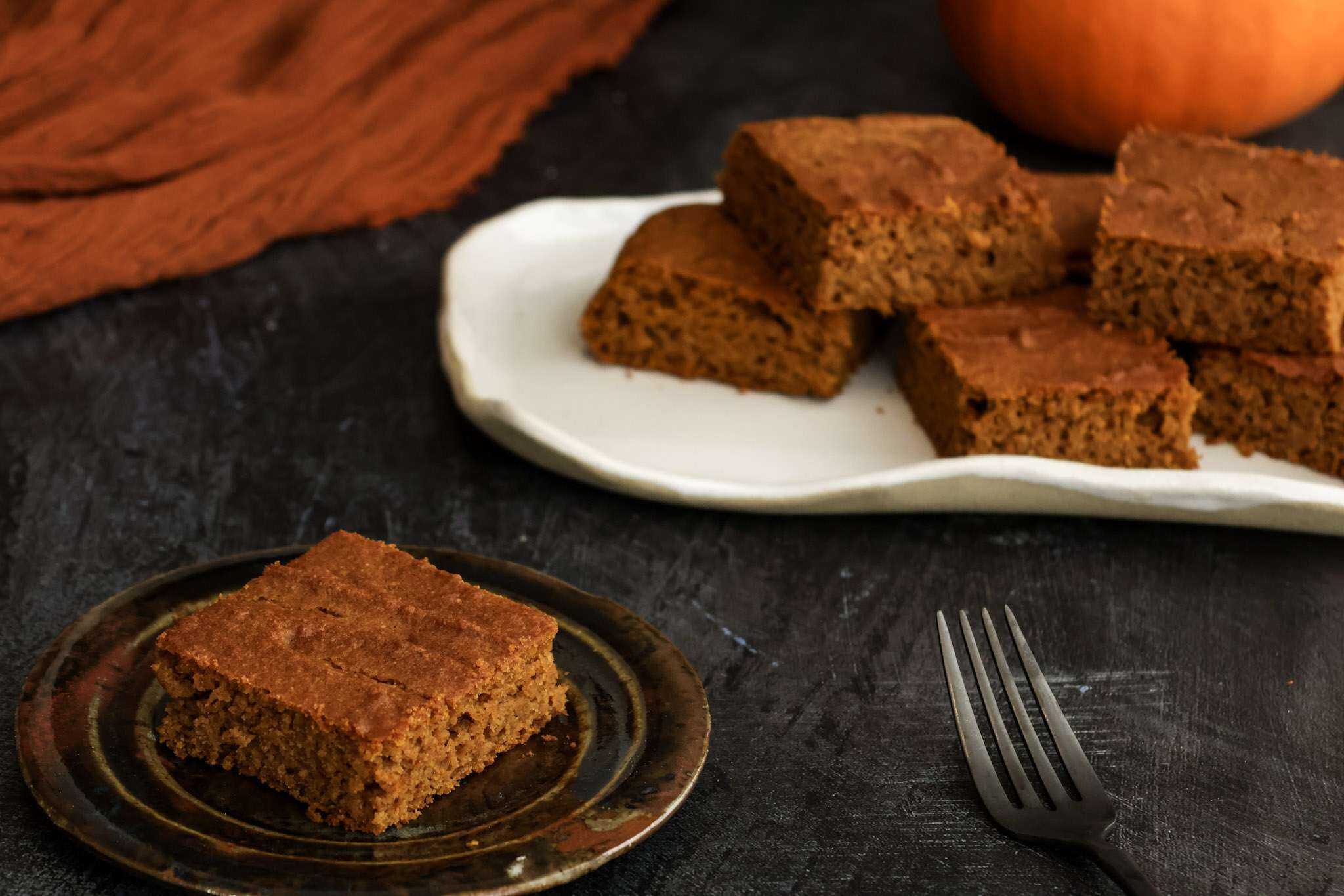 A small plate with a pumpkin bar in the front of the picture and a tray of additional bars is in the back - a fork, a partial mini pumpkin and an orange cloth can also be seen in the background.
