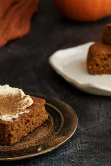 A small plate with a frosted pumpkin bar in the front of the picture and a tray of additional bars without frosting is in the back - a fork, a partial mini pumpkin and an orange cloth can also be seen in the background.
