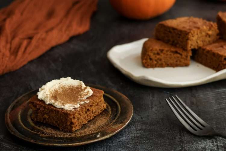 A small plate with a frosted pumpkin bar in the front of the picture and a tray of additional bars without frosting is in the back - a fork, a partial mini pumpkin and an orange cloth can also be seen in the background.