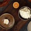 A bowl with orange mascarpone frosting, along with a plate with a frosted pumpkin bar and half an orange are displayed on a wooden board - next to the board is an orange cloth and an additional plate with a frosted pumpkin bar