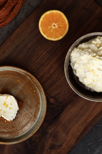 A bowl with orange mascarpone frosting, along with a plate with a frosted pumpkin bar and half an orange are displayed on a wooden board - next to the board is an orange cloth and an additional plate with a frosted pumpkin bar