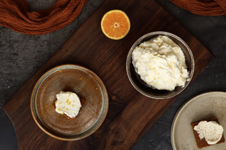 A bowl with orange mascarpone frosting, along with a plate with a frosted pumpkin bar and half an orange are displayed on a wooden board - next to the board is an orange cloth and an additional plate with a frosted pumpkin bar