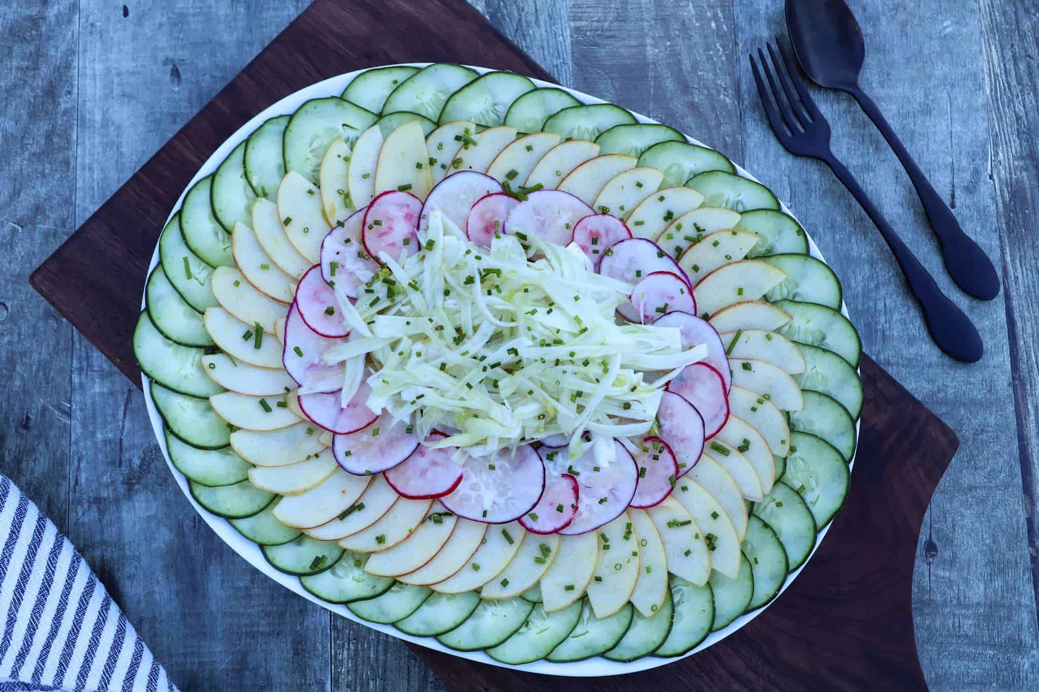 A plate of layered cucumber, apple, radish and fennel salad is placed on a wooden board with a napkin, spoon and fork in the background
