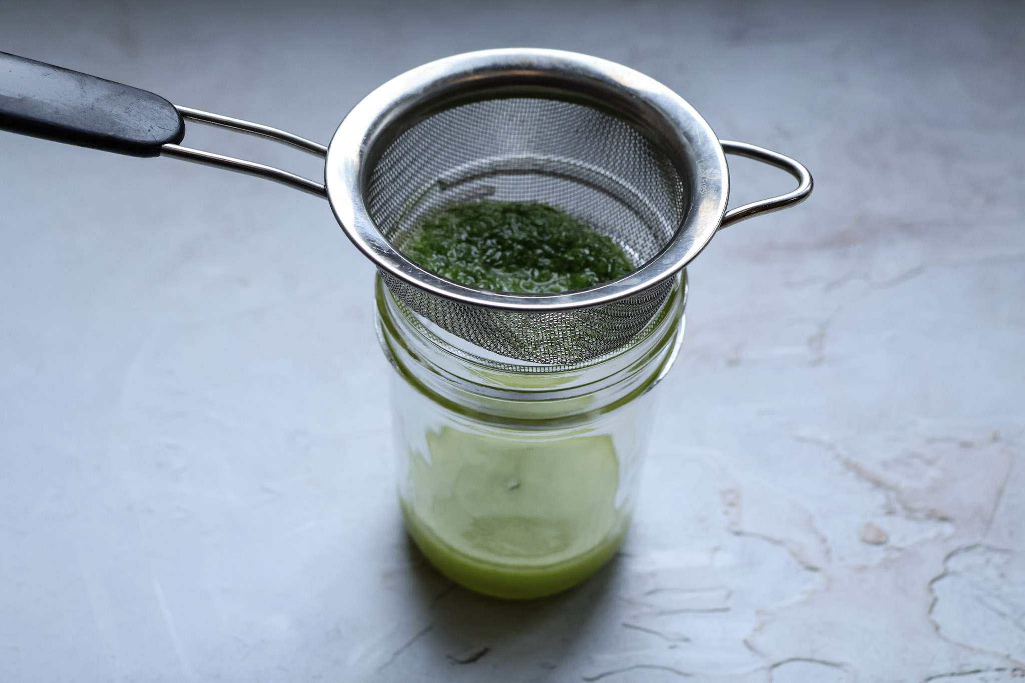 A small sieve on top of a small jar and herb oil is getting strained through it