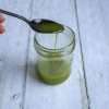 A small jar with herb oil and a hand holding a spoon over the top with additional oil drizzling into the jar