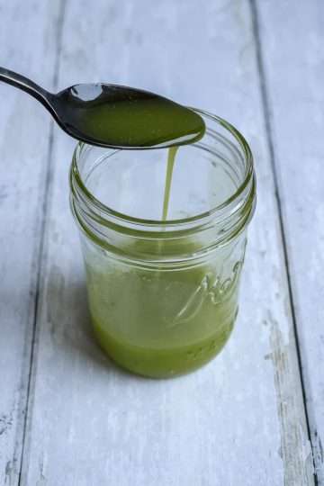A small jar with herb oil and a hand holding a spoon over the top with additional oil drizzling into the jar