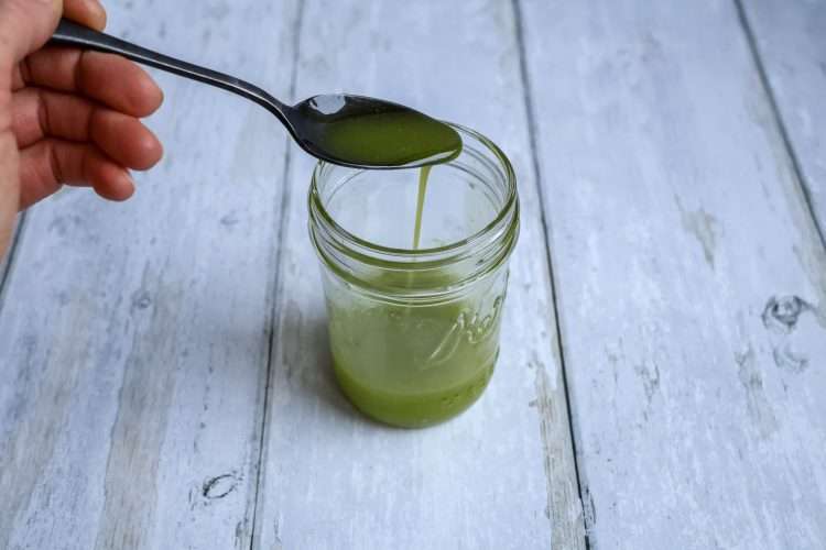A small jar with herb oil and a hand holding a spoon over the top with additional oil drizzling into the jar