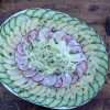 A plate of layered cucumber, apple, radish and fennel salad is placed on a wooden board with a napkin, spoon and fork in the background