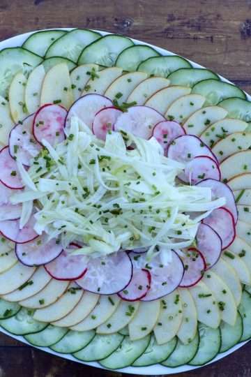 A plate of layered cucumber, apple, radish and fennel salad is placed on a wooden board with a napkin, spoon and fork in the background