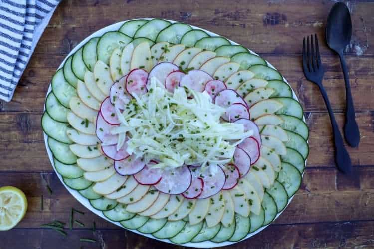 A plate of layered cucumber, apple, radish and fennel salad is placed on a wooden board with a napkin, spoon and fork in the background