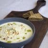 A wooden board with a bowl of spiced kohlrabi, fennel and apple soup with a drizzle of herb oil, sprinkle of hazelnuts, and a few diced apples and kohlrabi in the center. Two pieces of a skillet bread are in the background.