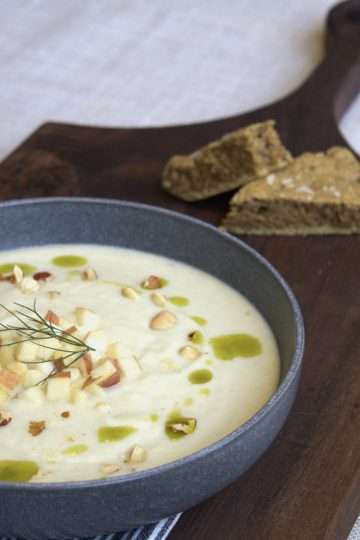 A wooden board with a bowl of spiced kohlrabi, fennel and apple soup with a drizzle of herb oil, sprinkle of hazelnuts, and a few diced apples and kohlrabi in the center. Two pieces of a skillet bread are in the background.