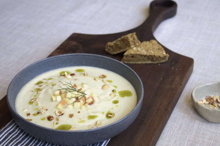 A wooden board with a bowl of spiced kohlrabi, fennel and apple soup with a drizzle of herb oil, sprinkle of hazelnuts, and a few diced apples and kohlrabi in the center. Two pieces of a skillet bread are in the background.