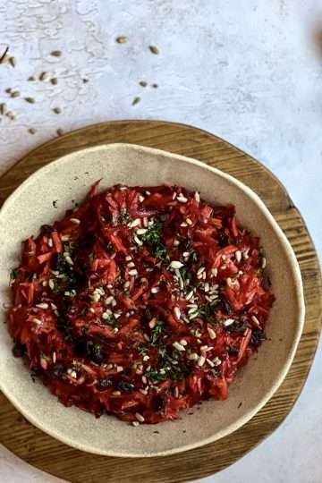 A bowl with crunchy beet and carrot slaw with apples is in the center, placed on top of a wooden board. A spoon and fork, as well as a beet and some sunflower seeds are in the background