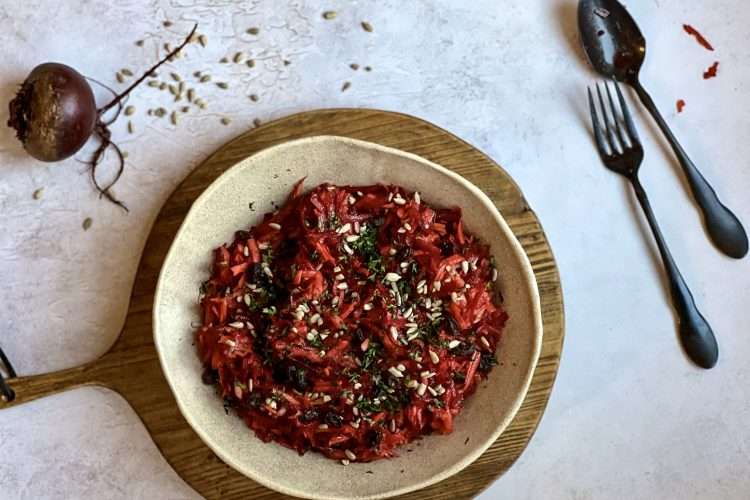A bowl with crunchy beet and carrot slaw with apples is in the center, placed on top of a wooden board. A spoon and fork, as well as a beet and some sunflower seeds are in the background