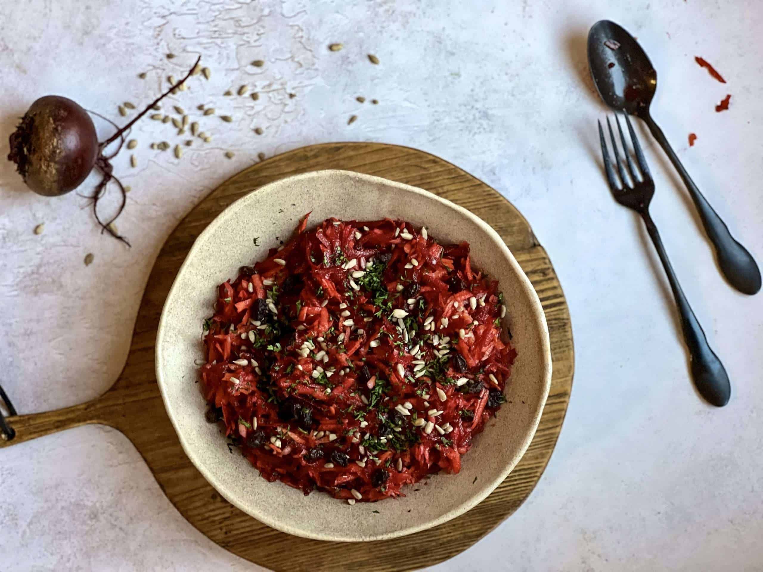 A bowl with crunchy beet and carrot slaw with apples is in the center, placed on top of a wooden board. A spoon and fork, as well as a beet and some sunflower seeds are in the background
