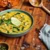 A green bowl holding creamy chicken and pumpkin chili that was garnished with two lime slices - a roasted pumpkin can be seen in the background, along with a wooden ladle, a green and white striped napkin and a small bowl of pumpkin seeds