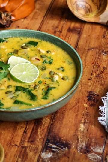 A green bowl holding creamy chicken and pumpkin chili that was garnished with two lime slices - a roasted pumpkin can be seen in the background, along with a wooden ladle, a green and white striped napkin and a small bowl of pumpkin seeds