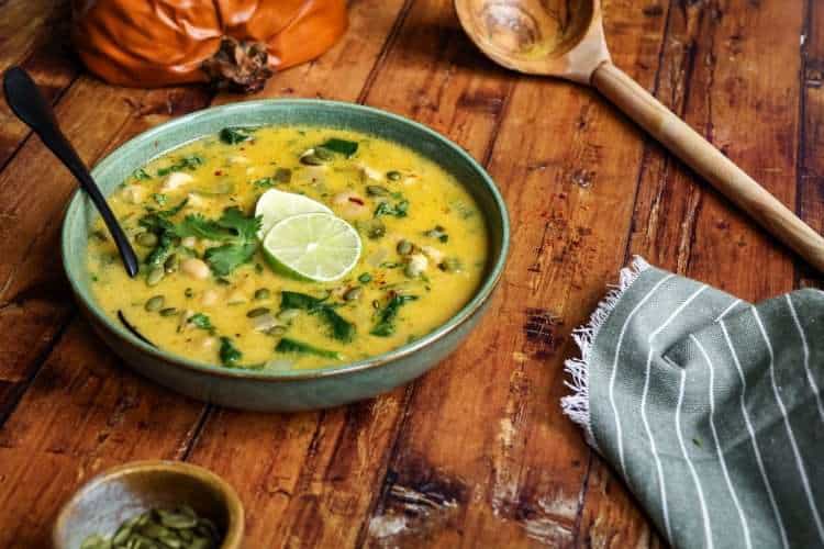A green bowl holding creamy chicken and pumpkin chili that was garnished with two lime slices - a roasted pumpkin can be seen in the background, along with a wooden ladle, a green and white striped napkin and a small bowl of pumpkin seeds