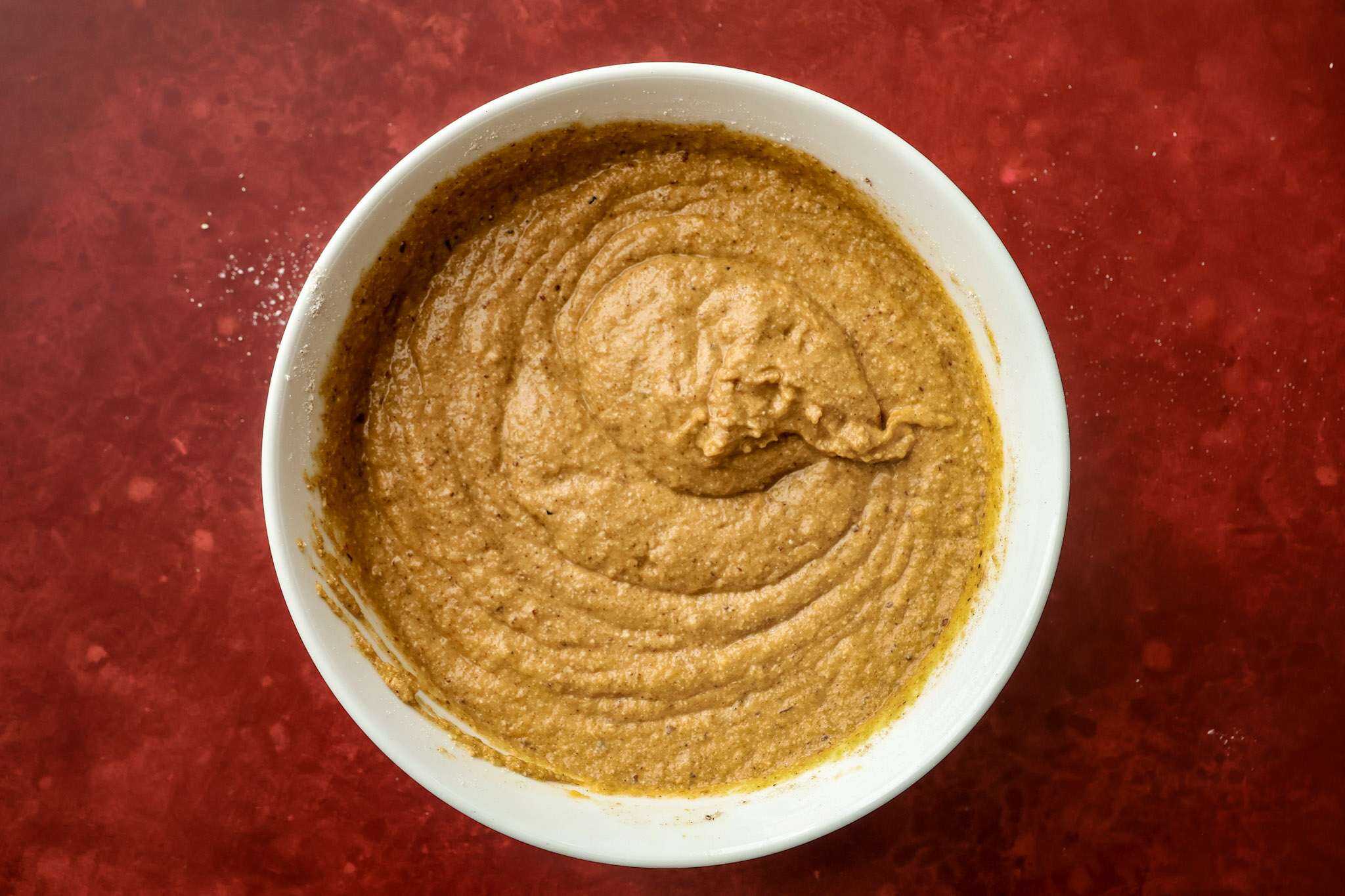 a white bowl on a red surface -the bowl contains the batter for tender gluten-free pumpkin muffins