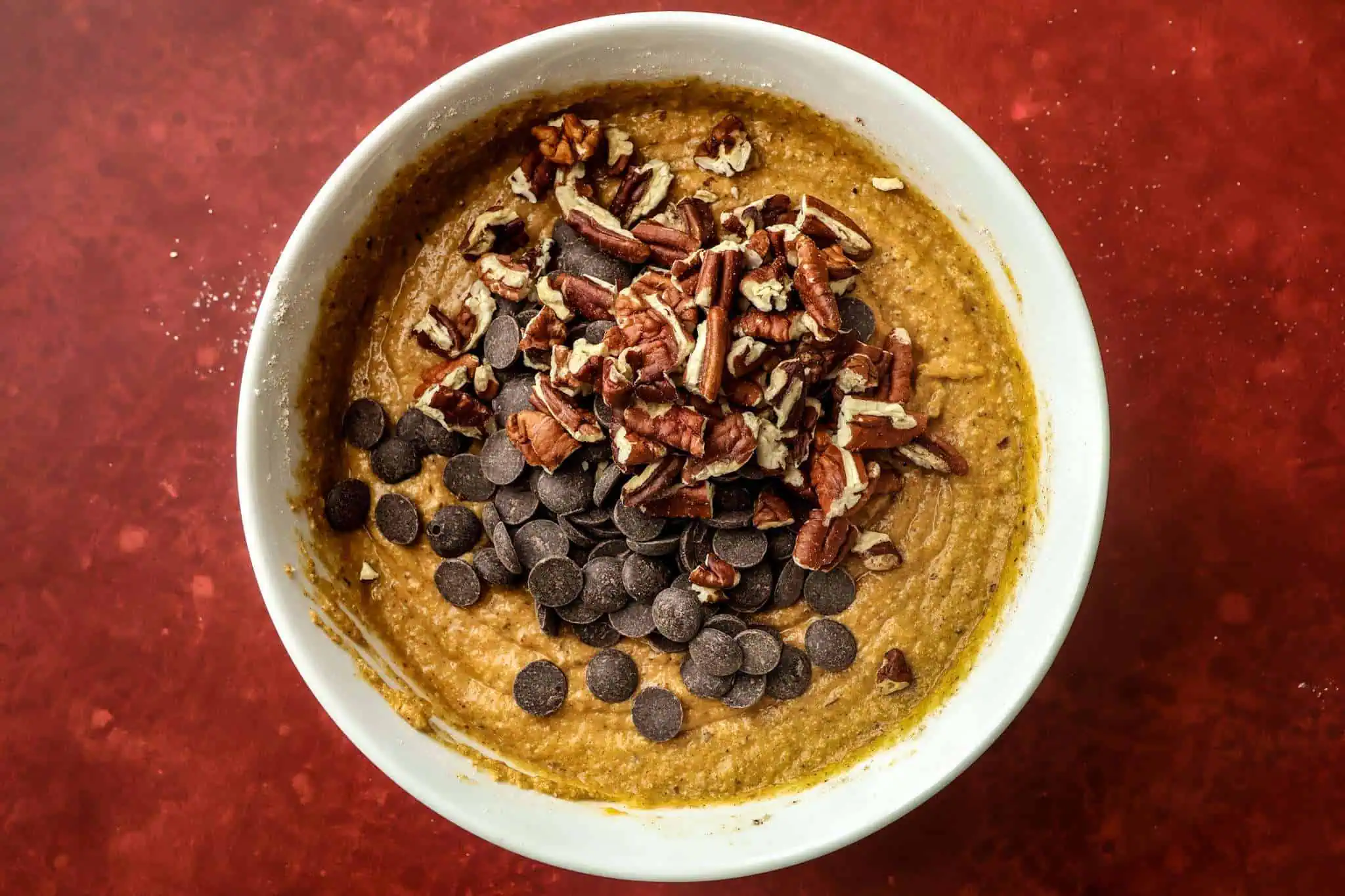 a white bowl on a red surface -the bowl contains the batter for tender gluten-free pumpkin muffins with chocolate chips and toasted pecans having just been added on top