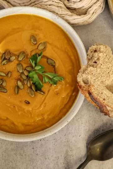a bowl of creamy winter vegetable soup garnished with toasted pumpkin seeds and parsley - a piece of sourdough bread is resting on the bowl and a black spoon, a small bowl of pumpkin seeds and a creamy colored napkin are surrounding the bowl - a second bowl of soup is partially displayed in the background