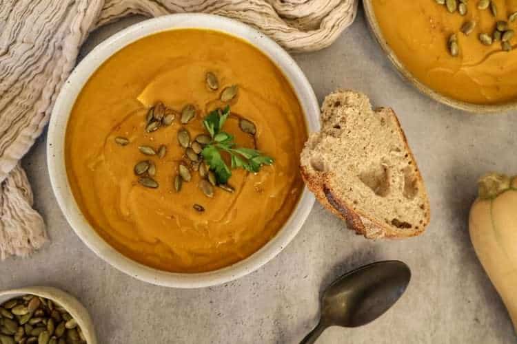 a bowl of creamy winter vegetable soup garnished with toasted pumpkin seeds and parsley - a piece of sourdough bread is resting on the bowl and a black spoon, a small bowl of pumpkin seeds and a creamy colored napkin are surrounding the bowl - a second bowl of soup is partially displayed in the background