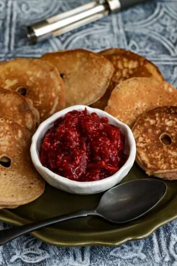 a green plate filled with apfelringen, a small while bowl of cranberry chia jam and a black spoon resting on the plate - an apple corer, a red cloth and an apple can be seen in the background
