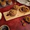 A deep red surface - on top is a small wooden board with two whole pumpkin muffins and one that has been loosened from the wrapper. One half is in the wrapper, the other half is next to the wooden board. A small bowl of chocolate chips can be partially seen, along with a small white pumpkin, a cream colored cloth in the background, a few scattered pecans and a cooling rack with additional muffins in the background