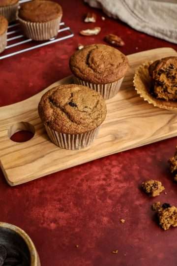 A deep red surface - on top is a small wooden board with two whole pumpkin muffins and one that has been loosened from the wrapper. One half is in the wrapper, the other half is next to the wooden board. A small bowl of chocolate chips can be partially seen, along with a small white pumpkin, a cream colored cloth in the background, a few scattered pecans and a cooling rack with additional muffins in the background