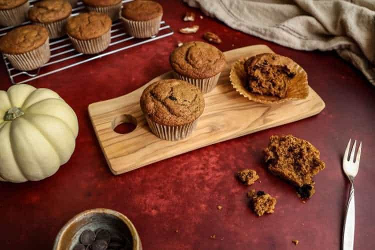 A deep red surface - on top is a small wooden board with two whole pumpkin muffins and one that has been loosened from the wrapper. One half is in the wrapper, the other half is next to the wooden board. A small bowl of chocolate chips can be partially seen, along with a small white pumpkin, a cream colored cloth in the background, a few scattered pecans and a cooling rack with additional muffins in the background
