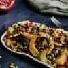 White rectangular platter filled with roasted delicata squash and fennel salad topped with pomegranate seeds, set on a dark grey background. A halved pomegranate and a small white bowl of pomegranate seeds sit nearby, with a fork, an olive-green cloth, and a few scattered pumpkin seeds in the background.